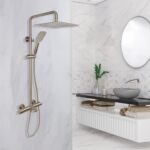 Modern bathroom with the Zen Thermostatic Shower Pipe System, wall-mounted rainfall showerhead and handheld, marble walls, round mirror, gray vessel sink, a vase with a plant, and neatly stacked towels.