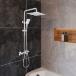Modern bathroom with the Zen Thermostatic Shower Pipe System, featuring a square rain showerhead and handheld spray, mounted on gray and black tiles. Above a white bathtub, a wooden shelf holds two bottles.