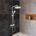 Modern bathroom with dark and light gray marble tiles, a bathtub, and the Zen Thermostatic Shower Pipe System featuring a large overhead shower and handheld sprayer. Two soap dispensers sit on a wooden shelf.