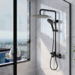 The Zen Thermostatic Shower Pipe System, featuring a black rain showerhead and handheld sprayer, is installed on a marble wall beside a large window overlooking the city skyline, tall buildings, and a river.