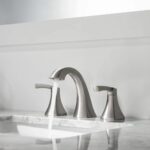 A Vermont 8 Inch Widespread Bathroom Sink Faucet in brushed nickel with two handles is installed on a white marble countertop, with water flowing from the spout into the sink.