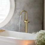 A modern bathroom showcases the Halo Floor Mount Tub Filler With Hand Shower in gold, dispensing water into a white tub. A round window and a vase with white flowers add to the tranquil ambiance.