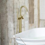 The Halo Floor Mount Tub Filler With Hand Shower, finished in gold, fills a white bathtub with water as rustic wooden wall panels add warmth to the background.