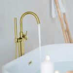 The Halo Floor Mount Tub Filler With Hand Shower fills a white tub, while a lit candle and towels on a wooden ladder in the background create a calm, spa-like atmosphere.