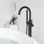 The Halo Floor Mount Tub Filler With Hand Shower in matte black features a curved spout beside a white bathtub in a modern, minimal bathroom, with a vanity and mirror visible in the background.