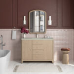A modern bathroom with a wooden vanity, marble countertop, arched mirror, twin wall lights, pink tile walls, a white bathtub, a round wooden stool, and a vase of purple flowers. A window is reflected in the mirror.