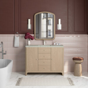A modern bathroom with a wooden vanity, marble countertop, arched mirror, twin wall lights, pink tile walls, a white bathtub, a round wooden stool, and a vase of purple flowers. A window is reflected in the mirror.