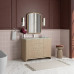 A modern bathroom with mauve walls, pink tile accents, a light wood vanity, an arched mirror, two vertical wall lights, a towel, a vase with flowers, a small wooden stool, and a fringed rug on a marble floor.