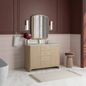 A modern bathroom with mauve walls, pink tile accents, a light wood vanity, an arched mirror, two vertical wall lights, a towel, a vase with flowers, a small wooden stool, and a fringed rug on a marble floor.