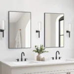 A modern bathroom showcases a double vanity with two Metal Framed Mirrors, black faucets, wall lights, a potted plant, and neatly stacked towels. In the background, a towel hangs on a gold hook.