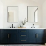Modern bathroom with a dark blue double sink vanity, gold handles, two black faucets, and Metal Framed Mirrors above each sink. A decorative tray holding a plant and toiletries sits on the counter.