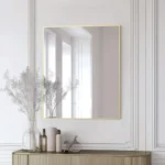 A modern Metal Framed Mirror with a thin gold edge hangs on a white paneled wall above a beige console table holding glass vases with dried flowers, bottles, and a small decorative bird statue.