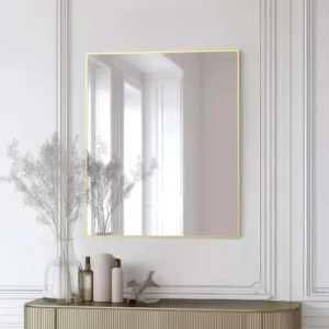 A modern Metal Framed Mirror with a thin gold edge hangs on a white paneled wall above a beige console table holding glass vases with dried flowers, bottles, and a small decorative bird statue.