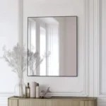 A Metal Framed Mirror with a slim black trim hangs on a white paneled wall above a console table featuring vases, dried plants, and bottles in a minimalist modern setting.