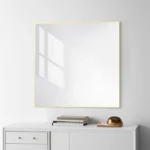 A Metal Framed Mirror with a thin gold edge hangs above a white cabinet on a white wall, reflecting sunlight and stacked books with a decorative gold object below.