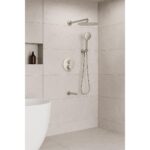 A modern bathroom with beige tiled walls showcases the Halo Pressure Balanced Wall Mount Shower System, featuring a round rainfall showerhead, handheld shower, built-in shelf with a bottle, grab bar, and white bathtub.
