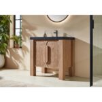 The Munich 36 Inch Bathroom Vanity with Top features two curved doors, a black countertop, and a modern faucet. It stands against a beige wall beneath a round mirror, near a window and potted plants.