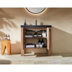 The Munich 36 Inch Bathroom Vanity with Top features open double doors displaying neatly organized towels and toiletries on shelves. The vanity's countertop holds soap dispensers, and a round mirror above reflects warm, streaming light.