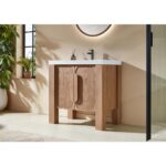 The Munich 36 Inch Bathroom Vanity with Top features a light wood finish, white countertop, and black faucet, paired with a round backlit mirror on a beige wall and a green plant by the window.