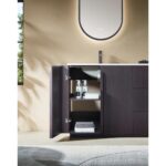 The Parma 48 Inch Bathroom Vanity With Top features dark wood cabinets and a door opening to shelves with towels and bottles, set beneath a curved, softly backlit mirror. A window and part of a patterned rug are also visible.