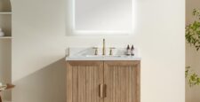 The Liora 30 Inch Bathroom Vanity features a light wood cabinet, white countertop, gold faucet, two small bottles, and a large illuminated rectangular mirror on a neutral wall, with shelves and a plant in the background.