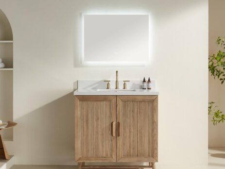 The Liora 30 Inch Bathroom Vanity features a light wood cabinet, white countertop, gold faucet, two small bottles, and a large illuminated rectangular mirror on a neutral wall, with shelves and a plant in the background.
