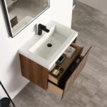 The Lille Wall Mounted Bathroom Vanity features a rectangular white sink, a black faucet, and two open wood drawers with toiletries and towels, set against a light gray tiled floor.
