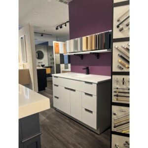 A showroom displays modern cabinets and countertops with sample cabinet doors and handles on the walls. The cabinetry is white with a sleek, minimalist design, and the wall behind features a deep purple accent color.