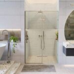 A modern bathroom features the Nova Hinged Frameless Shower Door, a white freestanding bathtub, terrazzo sink, wall plants, grey stone walls and flooring, and a gray mat in front of the shower.