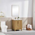 A modern bathroom featuring the Basel 24 Inch Bathroom Vanity with sink and mirror, a white toilet, towel basket, wall shelves with decor, and a neutral palette of beige paneling and a light gray rug.