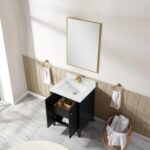A bathroom features the Torino 24 Inch Bathroom Vanity Cabinet in black, a white sink, gold faucet, wood paneling, a rectangular mirror above, two towels on gold rings, and a basket with rolled towels on the marble floor.