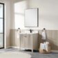 A bathroom with the Torino 30 Inch Bathroom Vanity Cabinet, topped by a framed mirror and black faucet. Towel rings flank the vanity, while a basket of rolled white towels sits beside it. Natural light pours in from a large window.