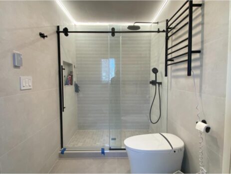 Modern bathroom with a glass sliding shower doorfeaturing a black frame, a wall-mounted showerhead, and a niche shelf. The room includes a white toilet, a heated towel rack, and light gray tiled walls.
