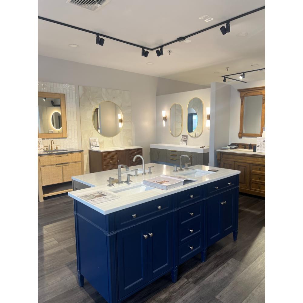 A showroom displays bathroom vanities, including a blue double-sink island with white countertops, wood and white vanities along the walls, round mirrors, and overhead track lighting.