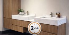 Modern bathroom with two wood and marble vanities, overhead cabinets, and dual sinks. Text promotes custom bathroom vanities ready in 2 weeks, local fabrication, made in USA, and white-glove delivery.