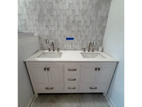 A white double-sink bathroom vanity with silver fixtures and four central drawers stands against a wall with glossy, rectangular gray tiles. The countertop is white, and the floor features large gray tiles.